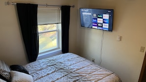 Et soveværelse med seng, et vindue med gardiner, et vægmonteret TV der viser streamingtjenester, og en loftlampe.