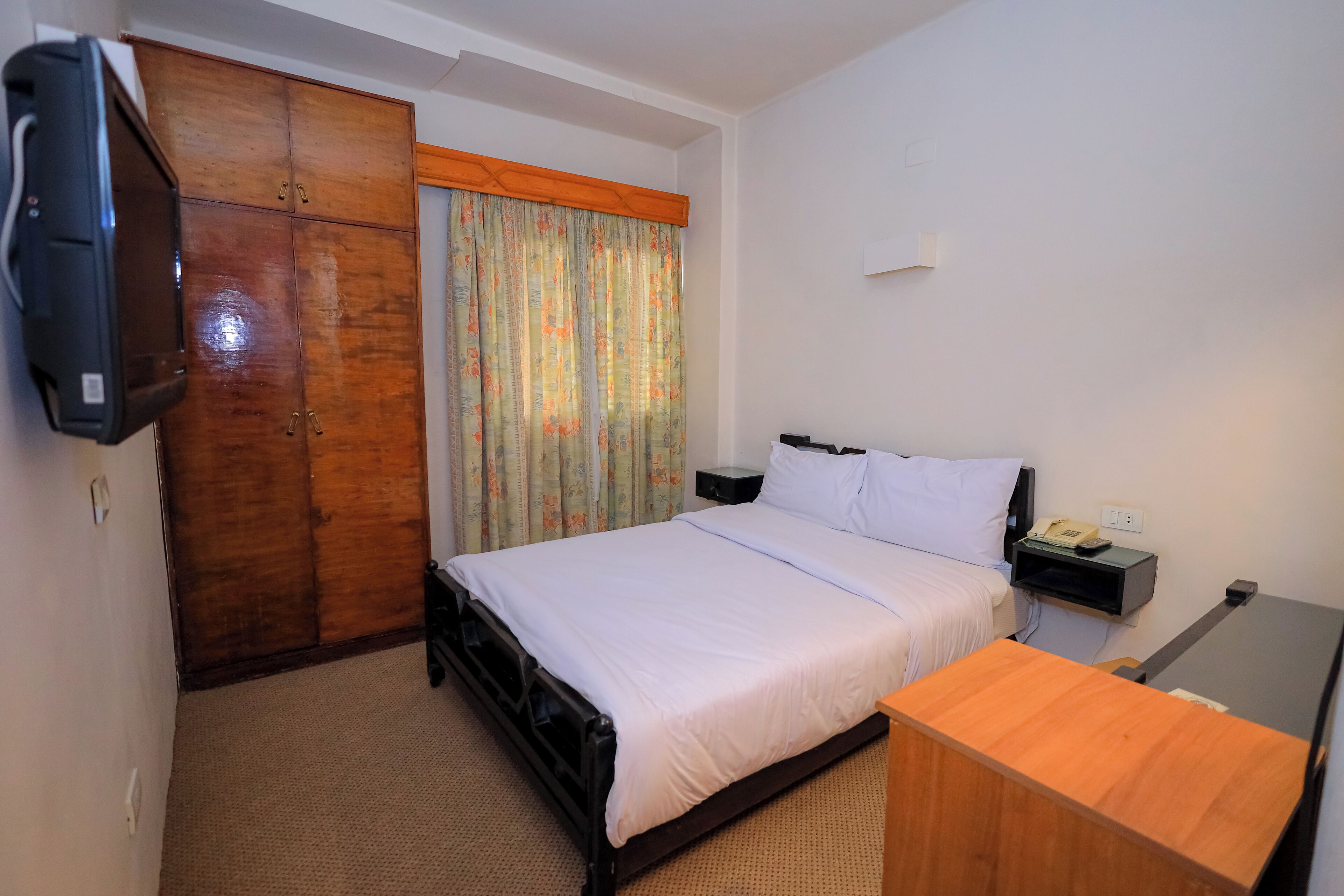 A hotel room with a bed, a wooden wardrobe, a television, a bedside table with a phone, and a curtain with a floral pattern.