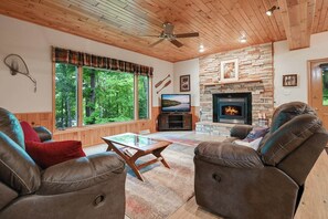 A cozy living room with a stone fireplace, leather sofas, a glass coffee table, and large windows offering a view of the forest.