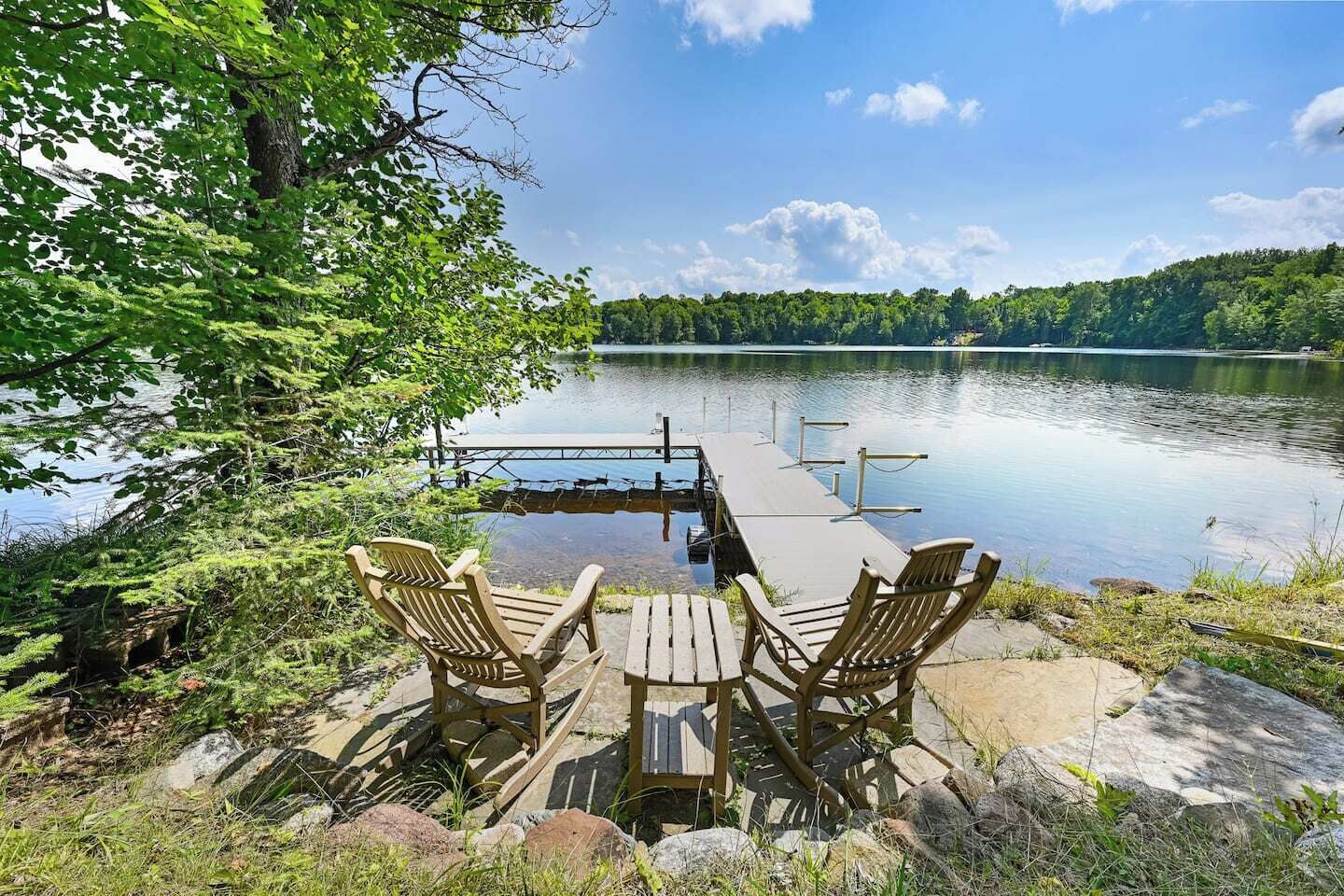 En terrass vid sjön med trämöbler, bestående av stolar och ett bord, med utsikt över en stilla sjö och skogbeklädda kullar.