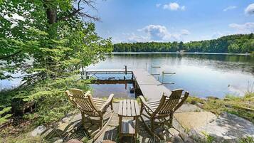 A lakeside deck with wooden chairs and a table, overlooking a calm lake and forested hills.