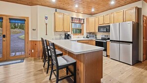 Una cocina moderna con armarios de madera, una isla grande con taburetes de bar, electrodomésticos de acero inoxidable y vistas al exterior a través de una puerta de vidrio.