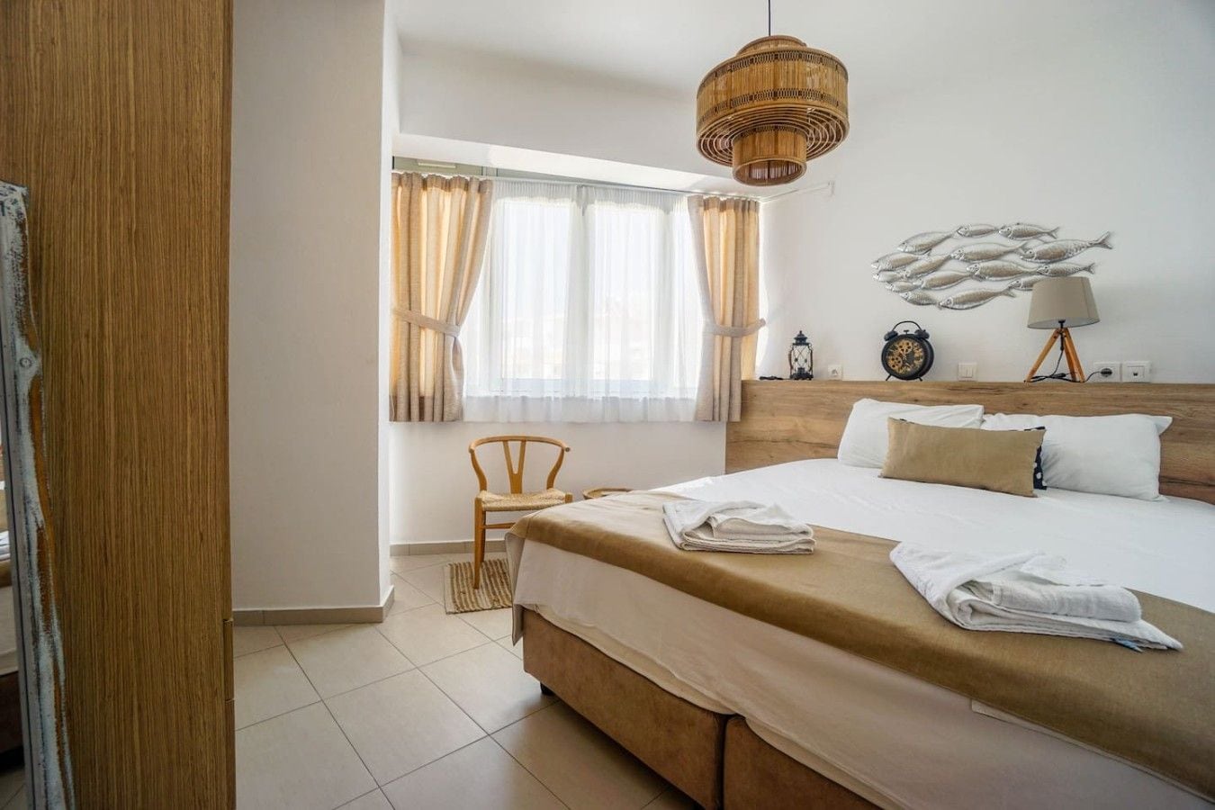 A hotel room with a large bed, a wooden headboard, a window with curtains, a bedside table with a lamp, and a wall clock.