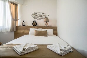 A neatly made bed with pillows and towels, a wall clock, a lamp, and a decorative fish wall art piece. - Urban beach sun(1c) The ultimate urban beach vibe. (Rethymno)