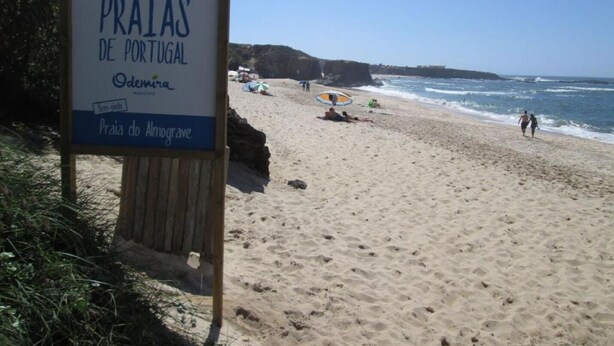 A beach sign with text in Portuguese, a sandy beach, and people in the background. - Almograve House (Almograve)