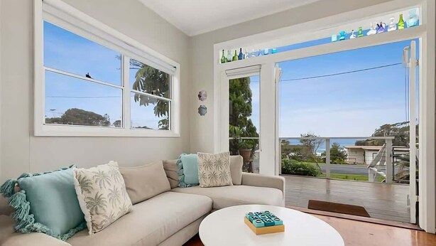 A living room with a beige sofa, a round coffee table, and a large sliding glass door leading to an outdoor area. - Lil Sea Shack by the Sea (Malua Bay)