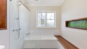 A bathroom with a shower, a bathtub, a window, and a wooden bench. - Lil Sea Shack by the Sea (Malua Bay)