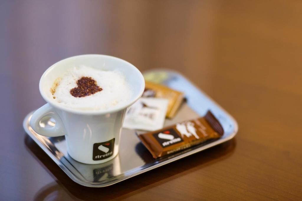 A cup of coffee with a heart-shaped design on the foam, a tray with a chocolate bar, and a small packet of creamer.