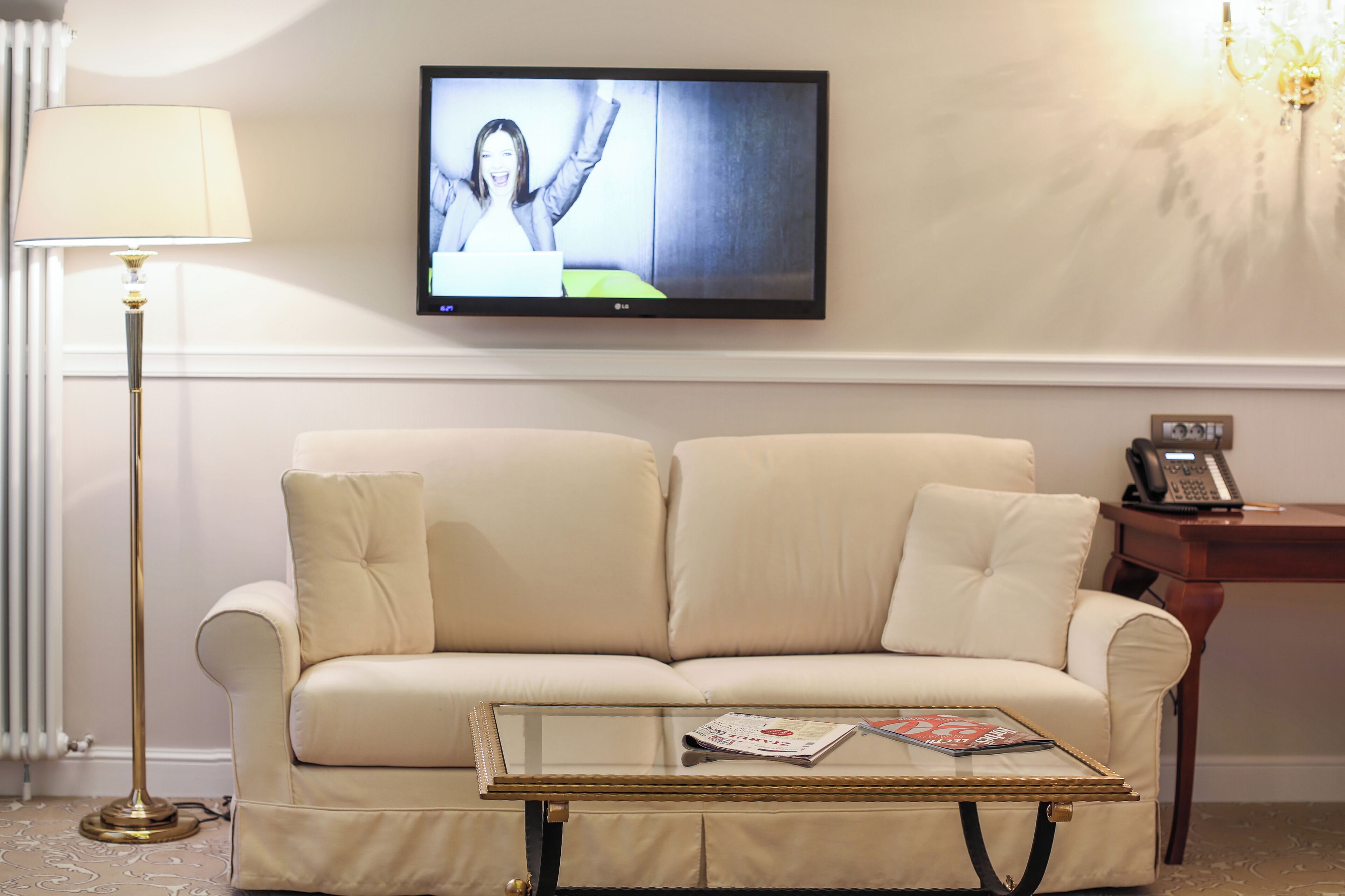 A living room with a beige sofa, a glass coffee table with magazines, a television on the wall, a floor lamp, and a telephone on a wooden side table.
