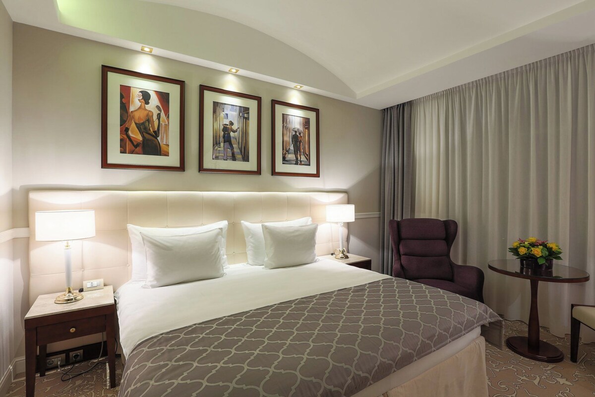 Een hotelkamer met een groot bed, twee nachtkastjes, een stoel, een klein tafeltje met bloemen en drie ingelijste kunstwerken aan de muur.