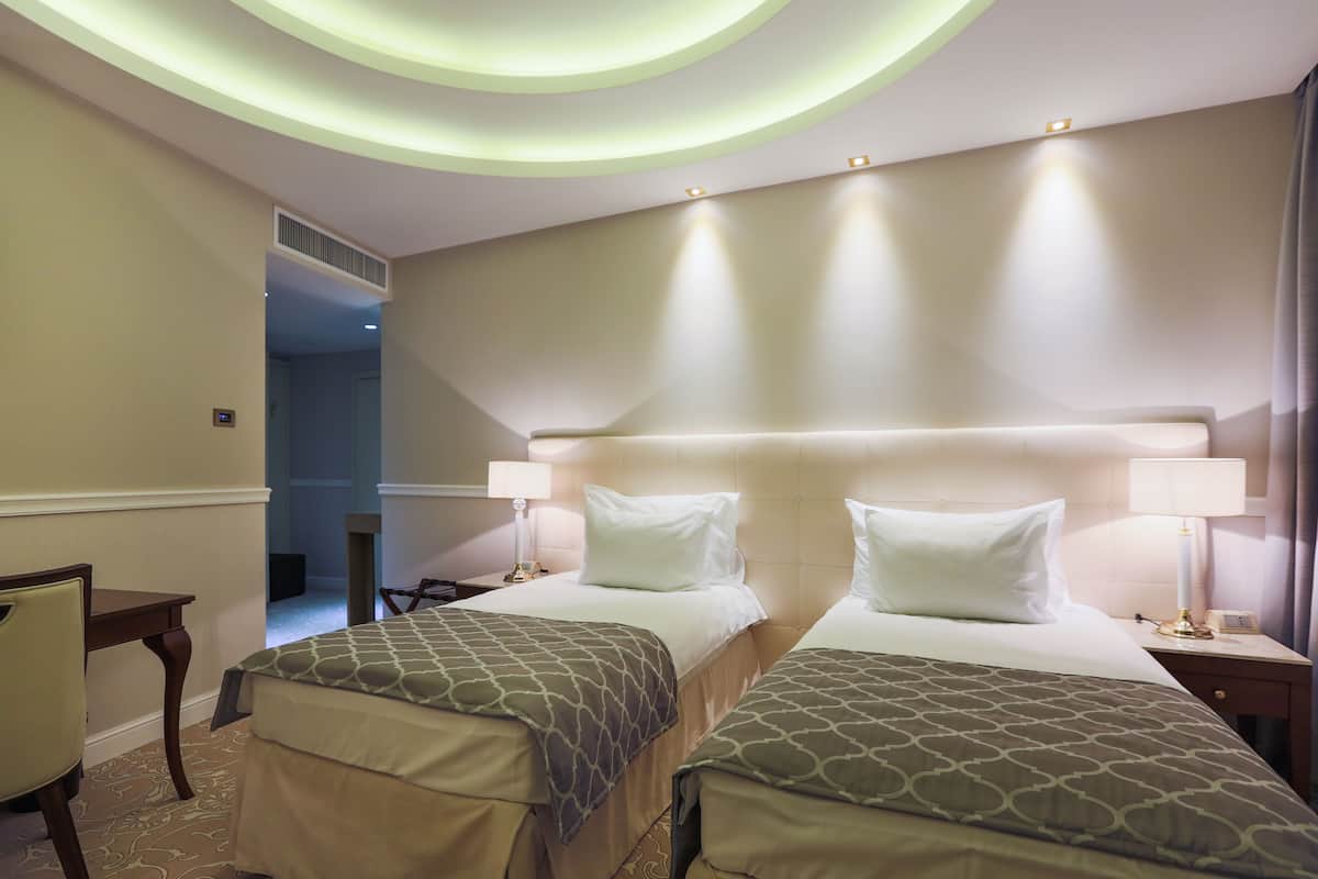 Een hotelkamer met twee bedden, een bureau en een stoel. Er is een plafondlamp en een deur naar een andere kamer.