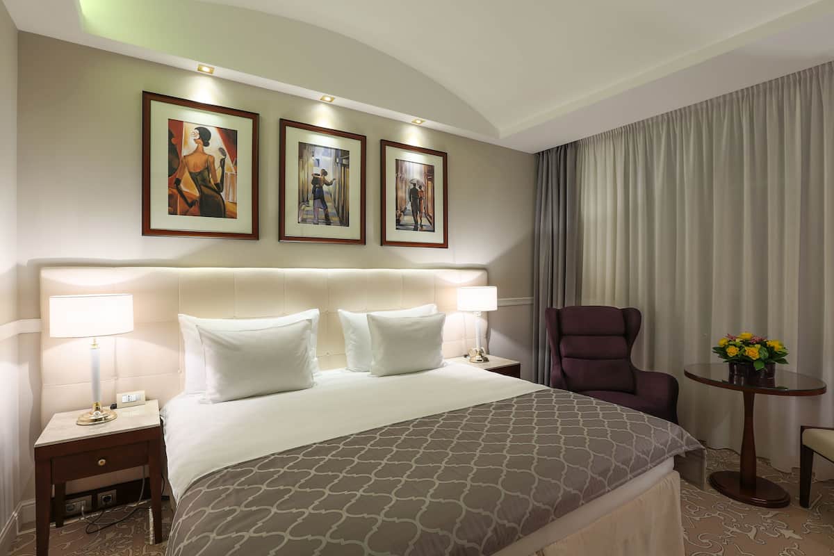 Een hotelkamer met een groot bed, twee bedlampen, een stoel, een bijzettafeltje met bloemen, en drie foto's in lijsten aan de muur.