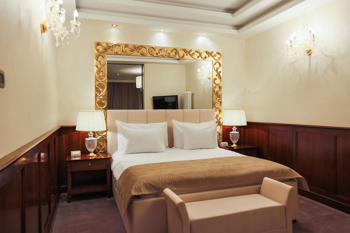Een hotelkamer met een groot bed, twee nachtkastjes met lampen, een grote spiegel met een sierlijk gouden frame, en een fauteuil.