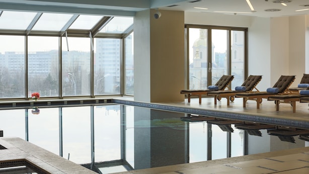 Une piscine intérieure avec des parois en verre transparent, des chaises longues et une vue sur les bâtiments environnants.