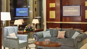 Un hall d’hôtel avec un canapé gris, des fauteuils, une table basse et un téléviseur affichant le nom de l’hôtel.
