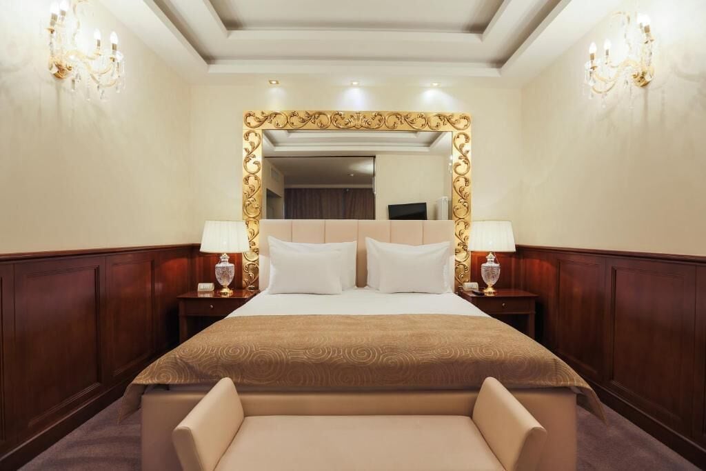 A hotel room with a large bed, two bedside tables with lamps, a mirror with an ornate frame, and a chandelier.