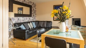 A modern living room with a black leather sofa, a glass coffee table, and a dining table with chairs. There is a vase with yellow flowers on the table, and a framed picture on the wall. - Amazing Castle View Apartment (Edinburgh)