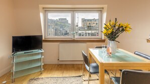 A modern living room with a television on a glass stand, a dining table with chairs, a vase with yellow flowers, and a view of a castle through a large window. - Amazing Castle View Apartment (Edinburgh)