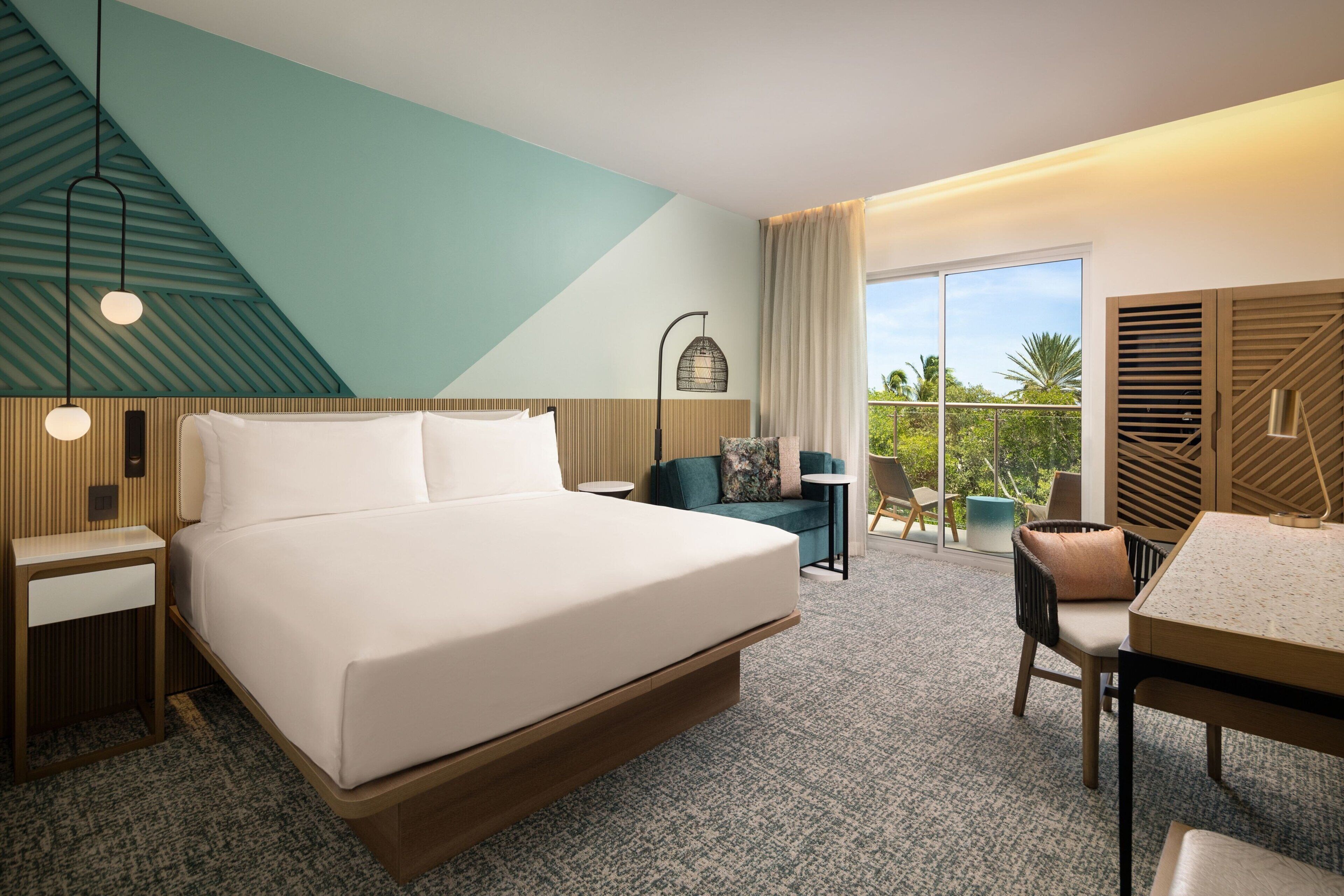 A modern hotel room with a large bed, a seating area with a table and chairs, and a balcony with a view of palm trees.