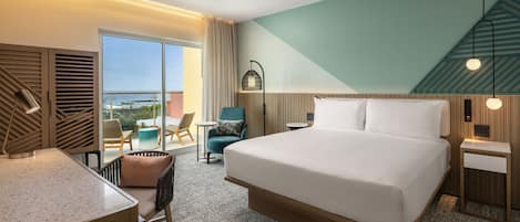A hotel room with a large bed, a desk, a chair, a small table, and a view of the ocean.
