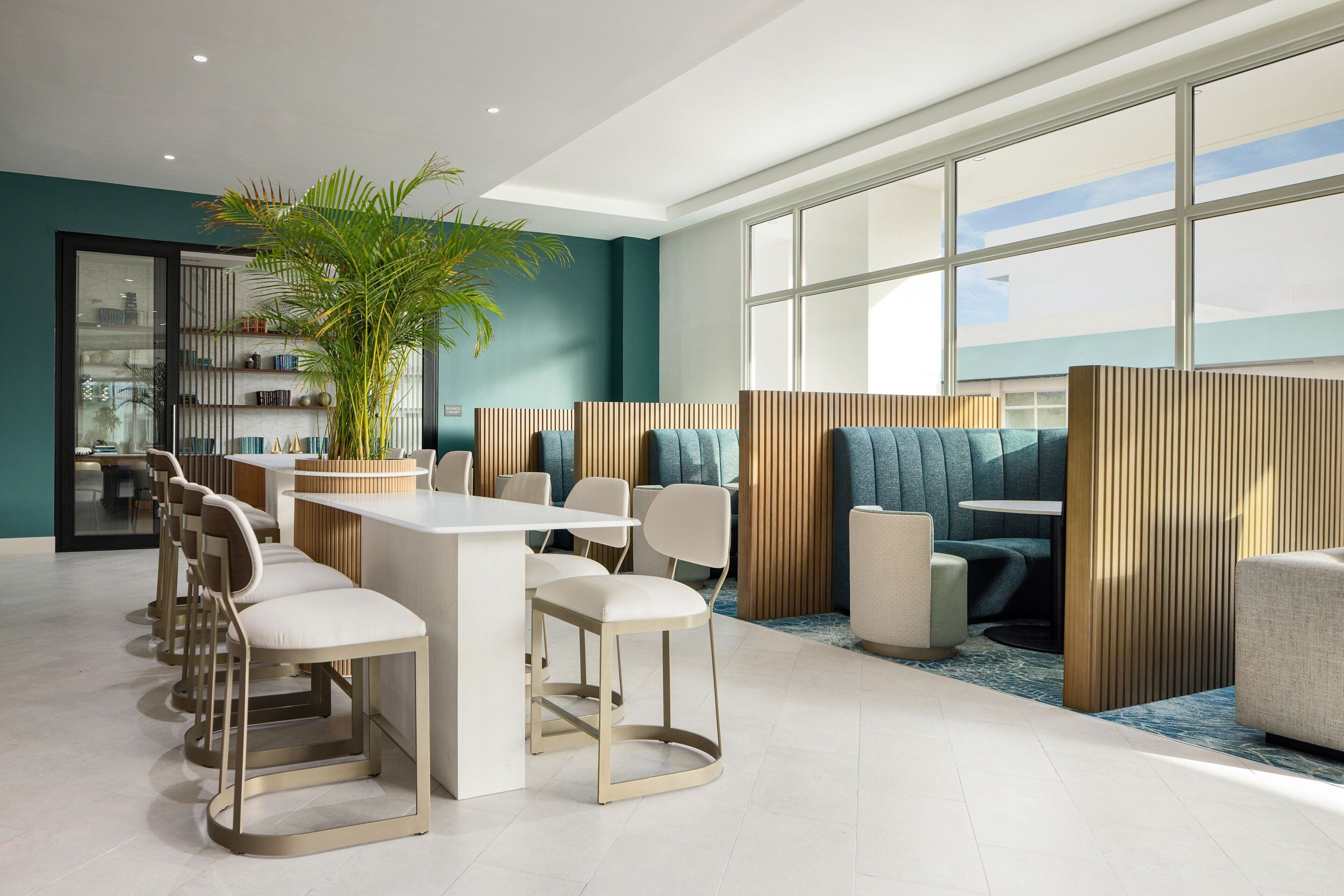 A modern dining area with a long table and chairs, a potted plant, and a view through large windows.