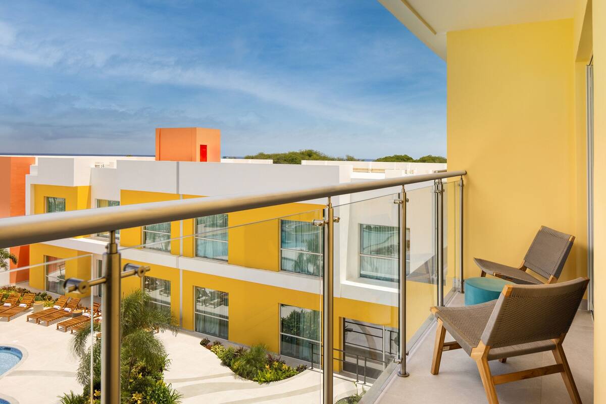 A modern hotel balcony with a glass railing, a pool, and lounge chairs.