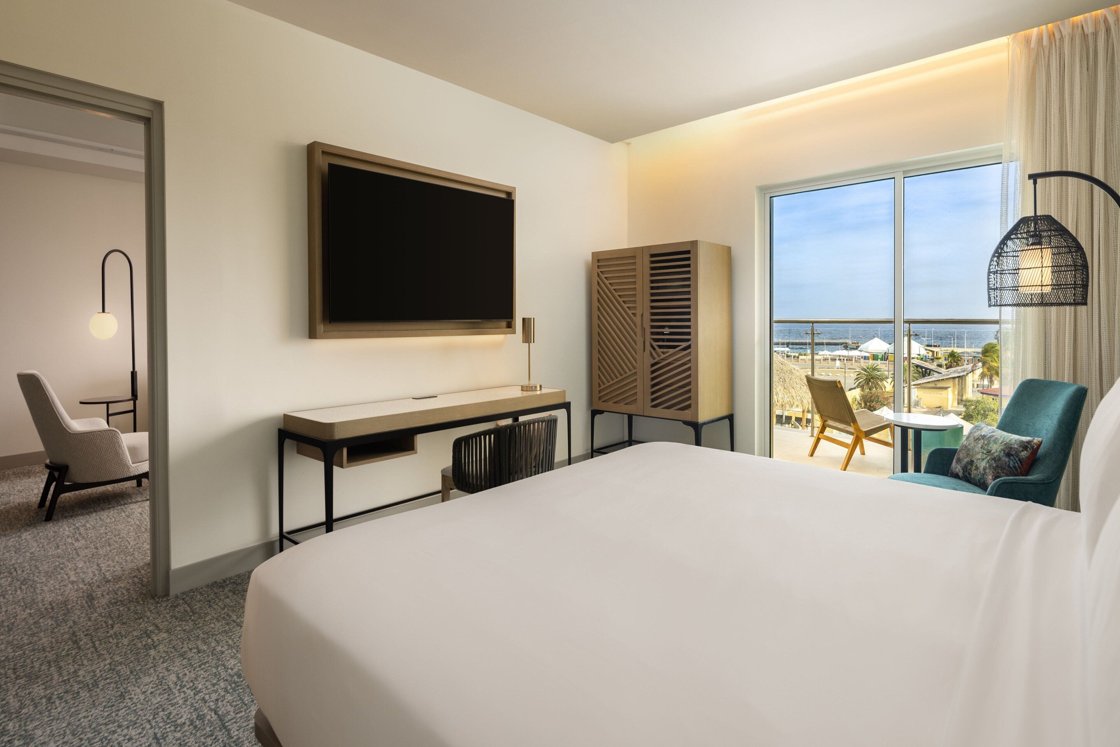 Habitación de hotel con una cama grande, un escritorio con silla, televisión, armario y balcón con vista a la playa.