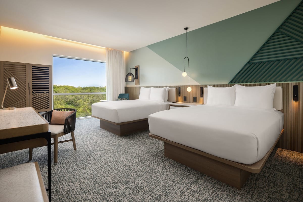 Ein Hotelzimmer mit zwei Betten, einem Schreibtisch und einem Stuhl. Ein großes Fenster bietet Blick auf eine grüne Umgebung.