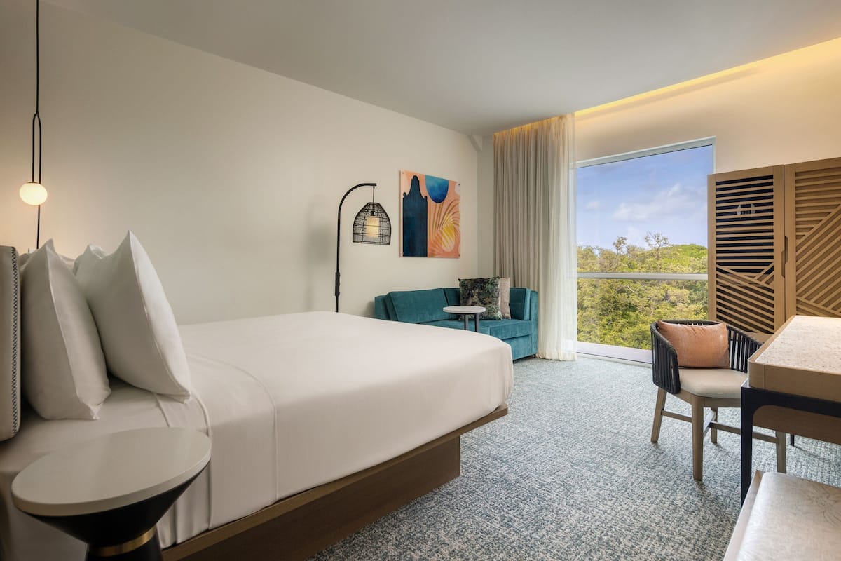 Ein Hotelzimmer mit einem großen Bett, einem kleinen runden Tisch, einer Couch, einem Sessel, einem Schreibtisch, einer Lampe und einem Gemälde.