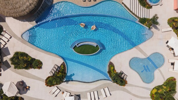 An aerial view of a uniquely shaped swimming pool with two people swimming, surrounded by lounge chairs and umbrellas. - Courtyard by Marriott Curacao (Willemstad)
