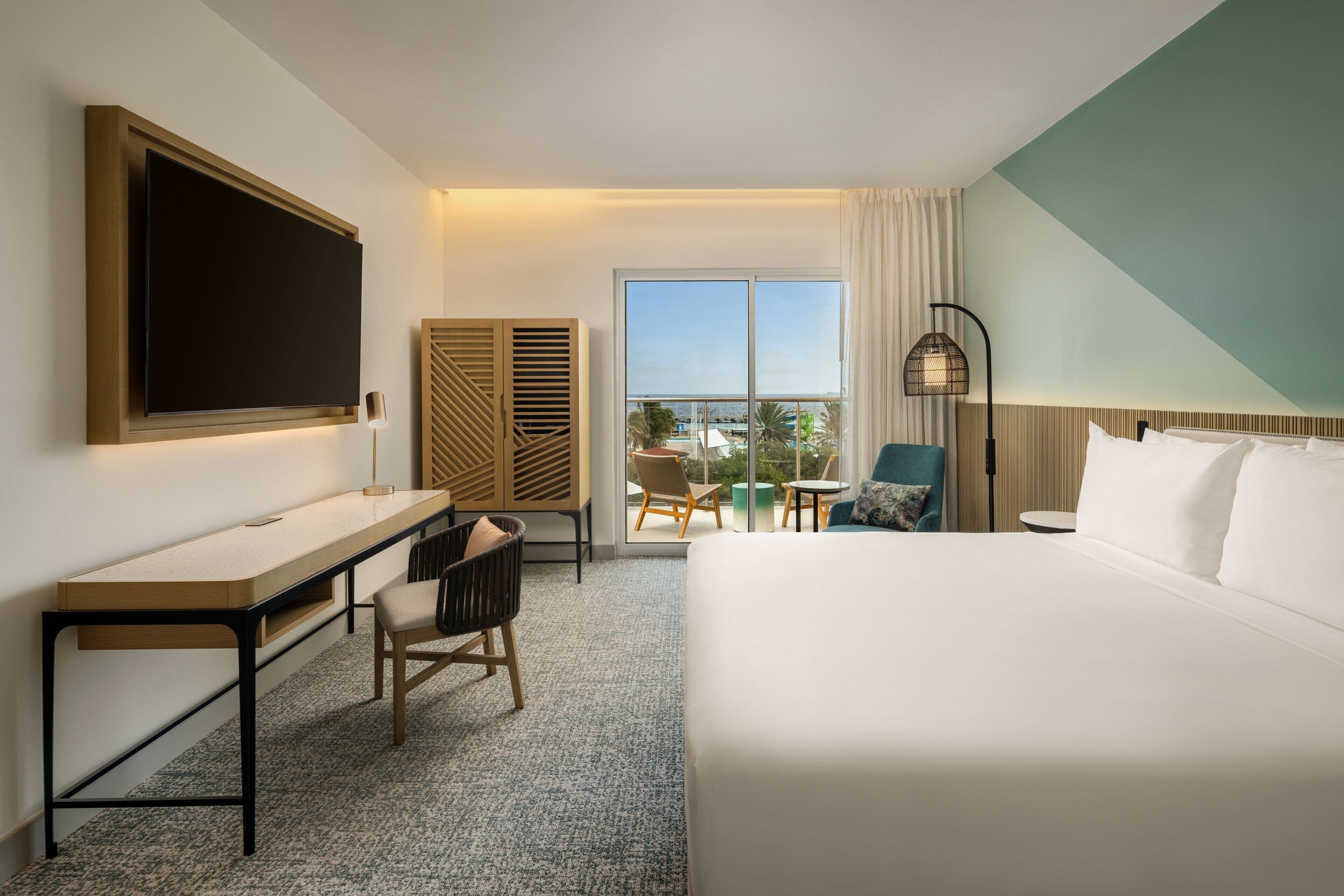 A hotel room with a bed, a desk, a chair, a TV, a window with a view, and a balcony with chairs.