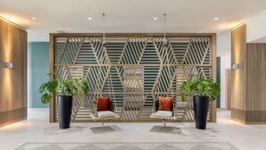 A modern lobby with a geometric wall design, two black potted plants, and two swing chairs with cushions. - Courtyard by Marriott Curacao (Willemstad)