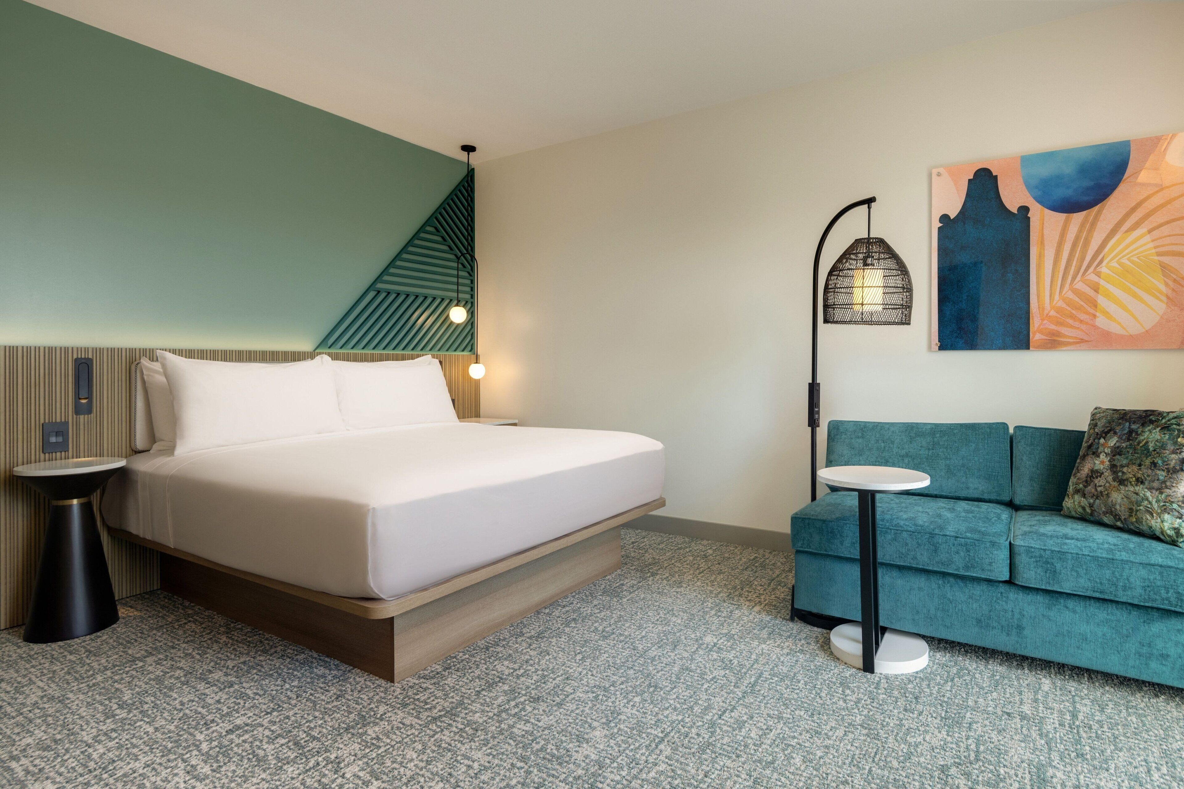 Une chambre d’hôtel avec un lit, un canapé, une petite table, une lampe fixée au mur et un tableau.