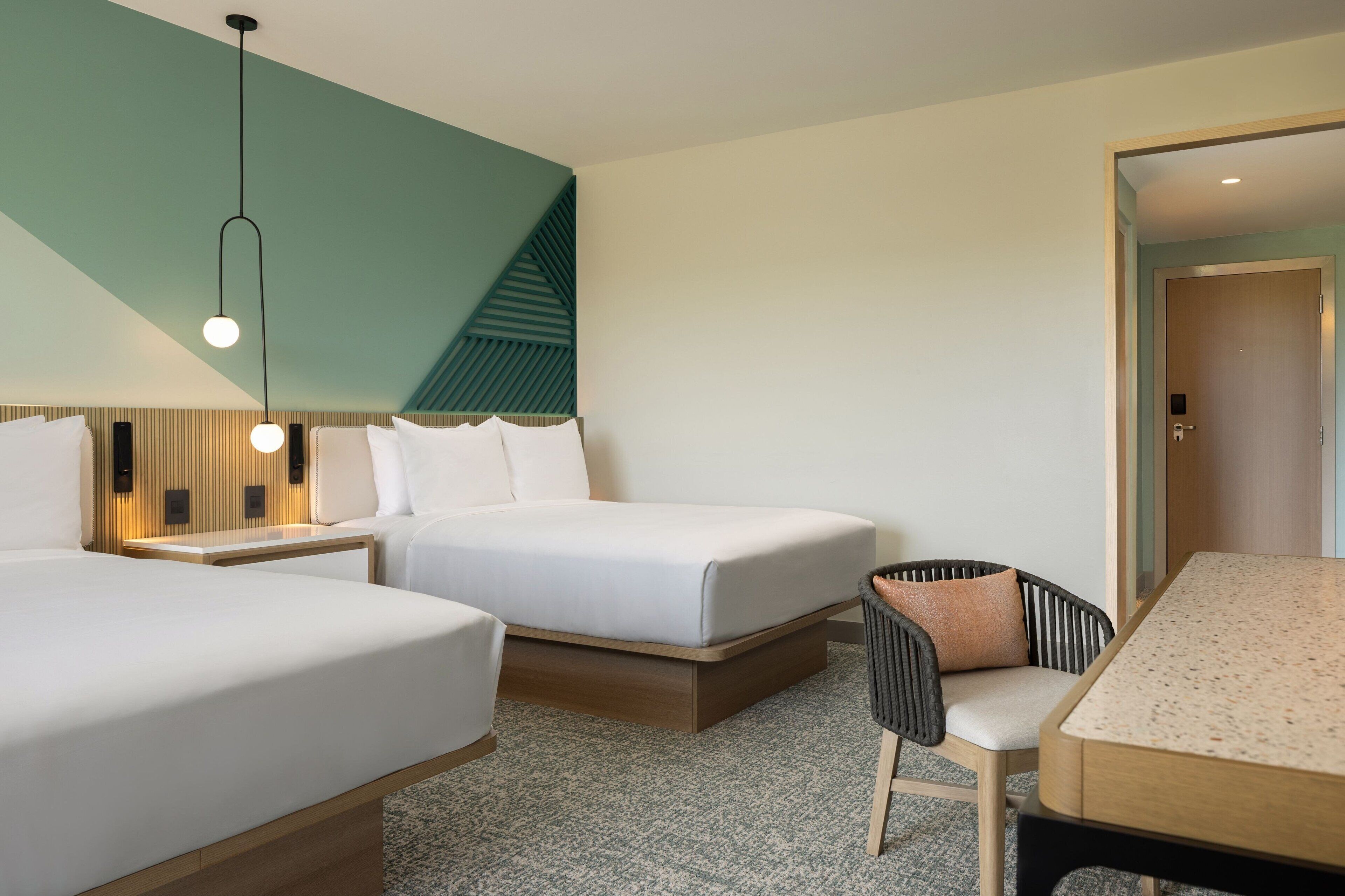 Quarto de hotel com duas camas, uma mesinha, uma cadeira e um espelho.