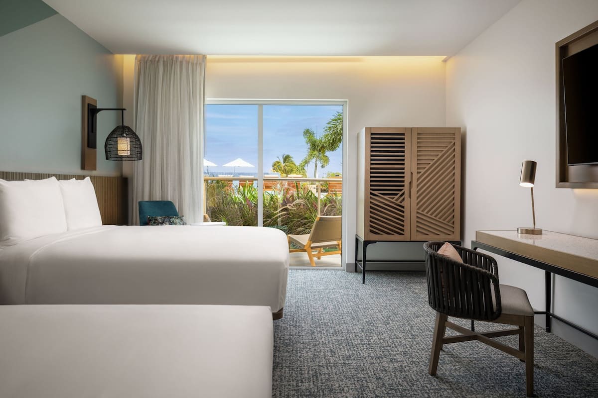 Ein Hotelzimmer mit zwei Betten, einem Schreibtisch, einem Stuhl, einem Fernseher und einem großen Fenster mit Blick auf Palmen und ein Gebäude.