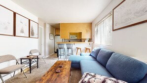 A modern living room with a blue sofa, a wooden coffee table, and a kitchen area with wooden cabinets and a microwave. - Bright Studio, On the Creek with a Pool On-site. Near Walking & Bike Trails (Mammoth Lakes)