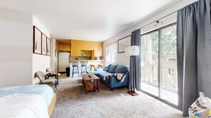 A compact hotel room with a kitchenette, a sofa, a dining table with chairs, and a large window with curtains.