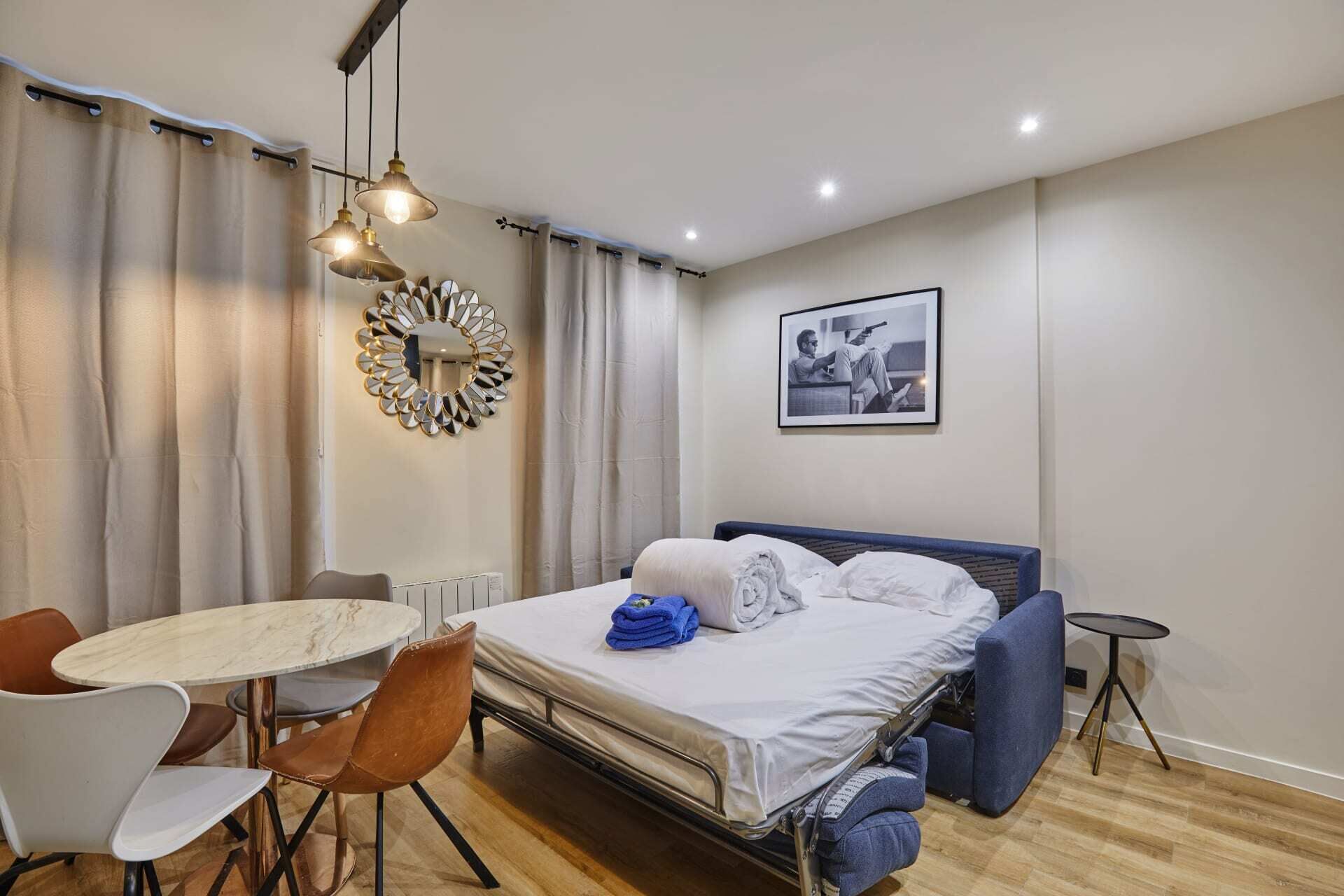Une chambre d’hôtel avec un lit, une table ronde entourée de chaises, une petite table ronde, un miroir et un tableau encadré accroché au mur.