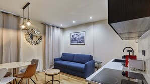 A compact living space with a dining area, a sofa, a small round table, a kitchen with a stove, and a wall-mounted mirror. - Meslay 6 - One Bedroom Apartment, Sleeps 4 (Paris)