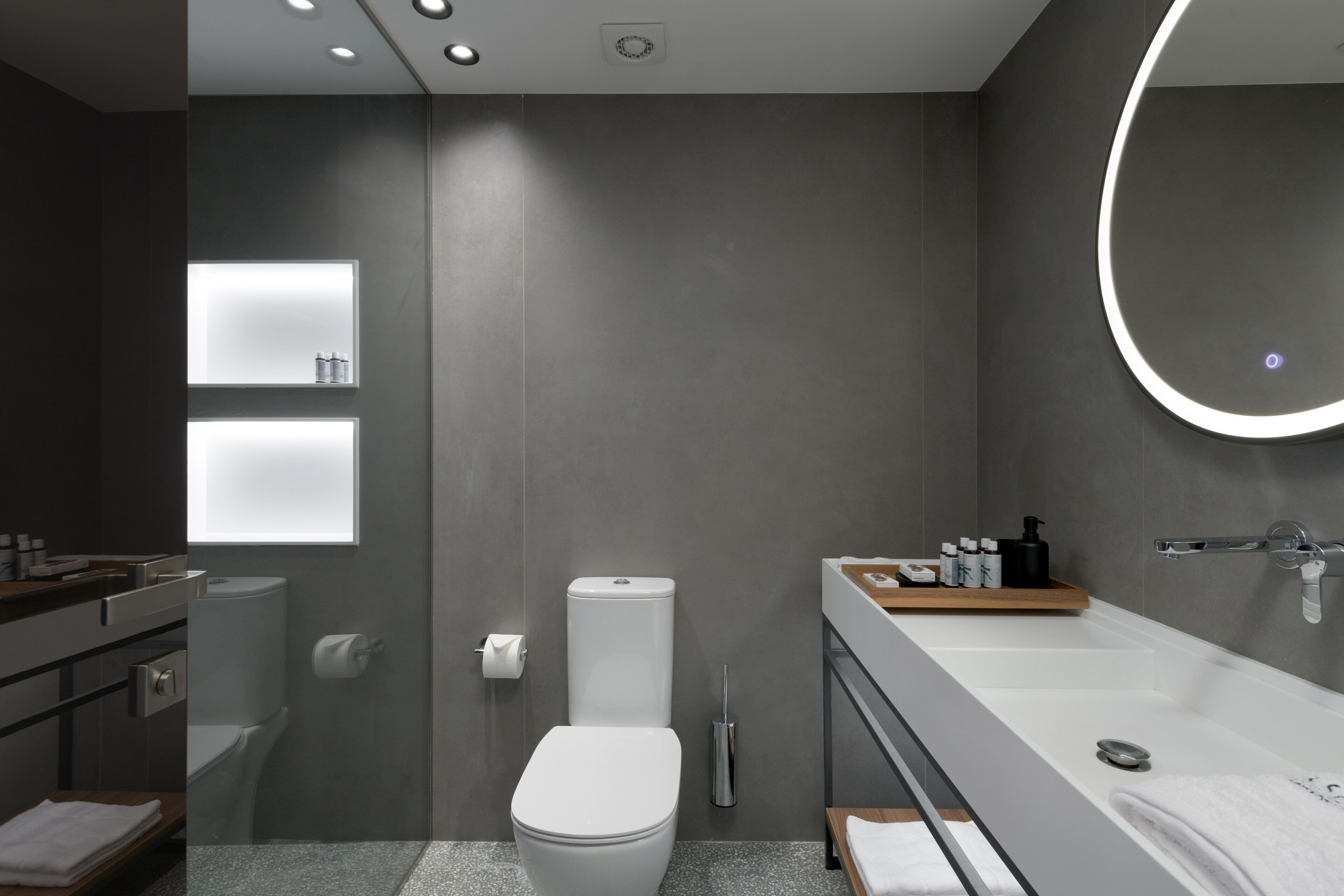 A modern bathroom with a toilet, sink, and bathtub. There are shelves with toiletries and a mirror with a light.