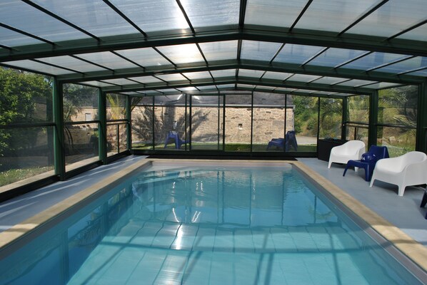 Piscina coberta com paredes de vidro transparente, cadeiras azuis e uma parede de pedra ao fundo.