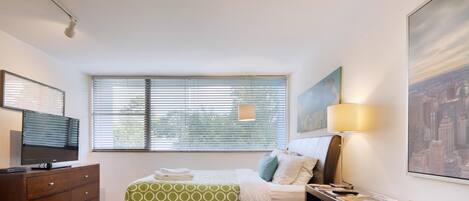 A hotel room with a bed, a desk, a lamp, a TV, and a window with blinds.