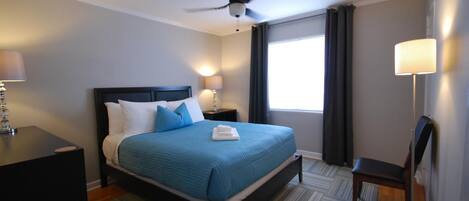 A hotel room with a bed, a ceiling fan, a nightstand with a lamp, a chair, and a window with curtains.