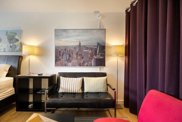 A modern hotel room with a black leather sofa, a red armchair, a cityscape painting, and two floor lamps.