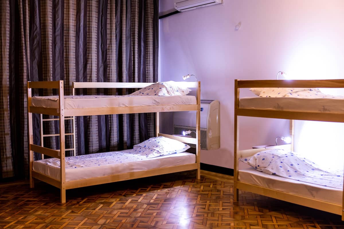 A room with two bunk beds, a wooden parquet floor, a window with curtains, and a wall-mounted air conditioner.
