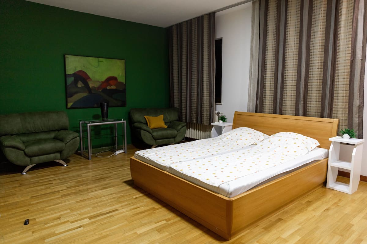 A bedroom with a wooden bed, green walls, a green armchair, a small white table, and a painting on the wall.