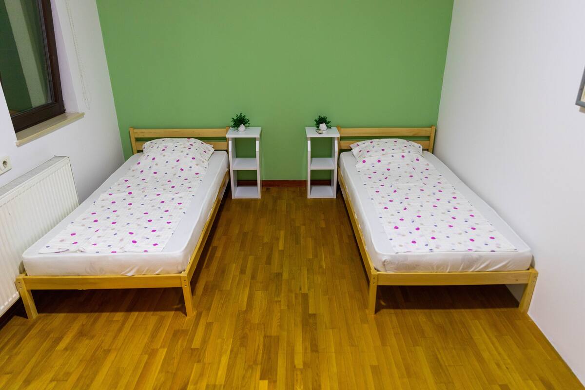 A room with two single beds, each with a white mattress and a colorful polka dot duvet. There are small white nightstands with plants on them. The room has a wooden floor and green walls.