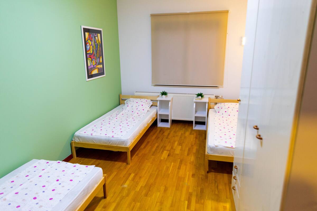 A room with two single beds, a radiator, a window with blinds, and a small table with a plant.