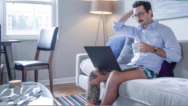 Een persoon zit op een bank met een laptop in de hand, een mok vasthoudend, in een kamer met een lamp, een stoel en een raam.