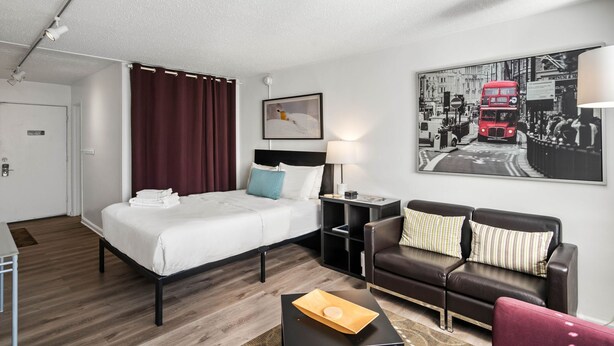 A modern hotel room with a bed, a desk, a sofa, and a large cityscape painting on the wall. - CCL Professional Studio w Wi-fi Perfect for Business Travelers (Atlanta)